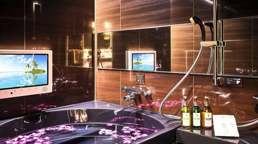 Dark jetted bathtub scattered with orchid petals beside a bathroom TV at Hotel Balian Resort Namba Shinsaibashi