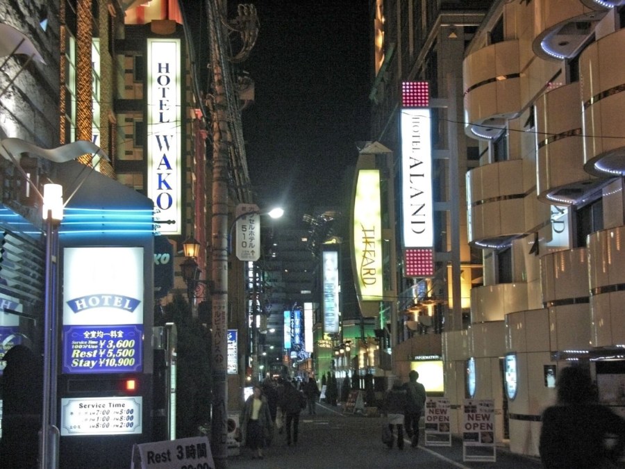 A street of love hotels at night with lit hotel signs and a board listing Rest and Stay prices